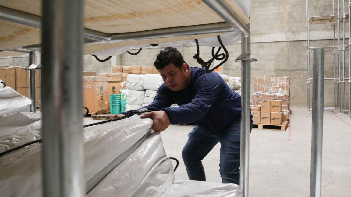 A man pulls a large white package from a shelf. He is in a warehouse. He is wearing jeans and a dark blue long sleeved top.