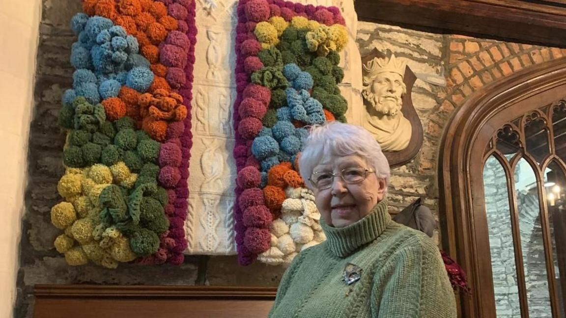 Volunteer Liz Fielding standing in front of  a knitted cross