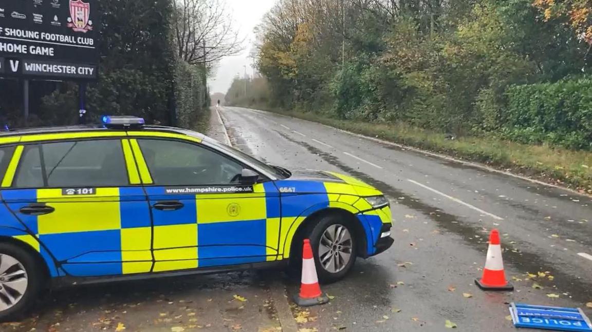 A police car is parked at the side of a road near some traffic cones. A 'police incident' sign has fallen over.