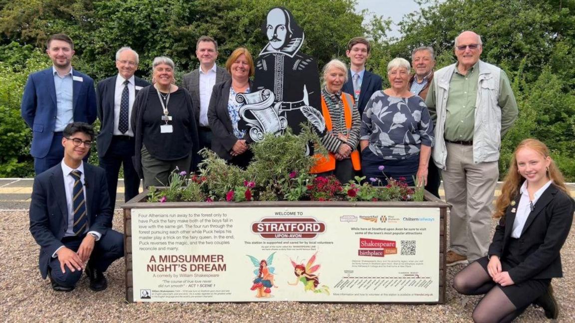 A group of people standing on a train platform behind a raised bed with bedding plants in and a black and white statue of William Shakespeare in the middle.