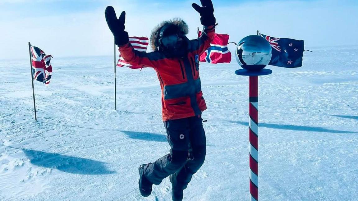 Cat Burford jumps for joy  as she arrives at the South Pole. She is dressed in a red jacket with hood and dark trousers and mittens and is surrounded by flags including the Union Jack and a glass sphere on top of a red and white painted pole.