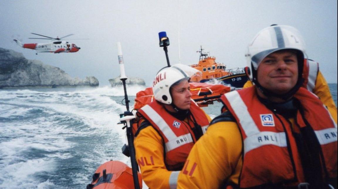 Nick Hayward in a lifeboat on the water with a colleague, and a search helicopter in the background.