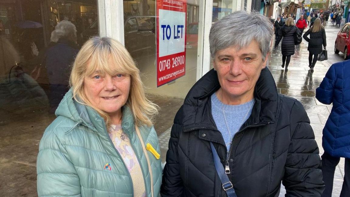 Linda on the left with blonde hair is wearing a green coat and Christine on the right has grey hair and is wearing a blue coat. People behind them on the pavement are walking.