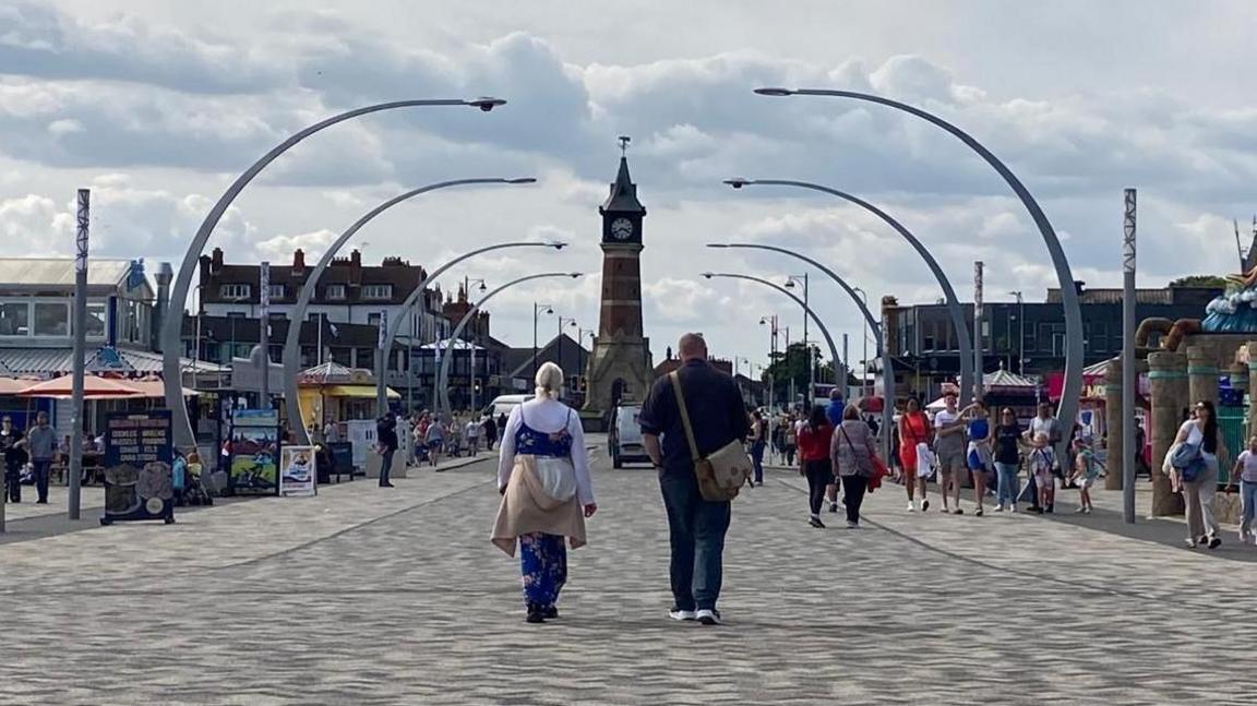 A pedestrian area of Skegness showing people walking and the town's clock tower in the distance. Stalls and rides are visible on either side of the stone walkway which also has large, curved street lights.