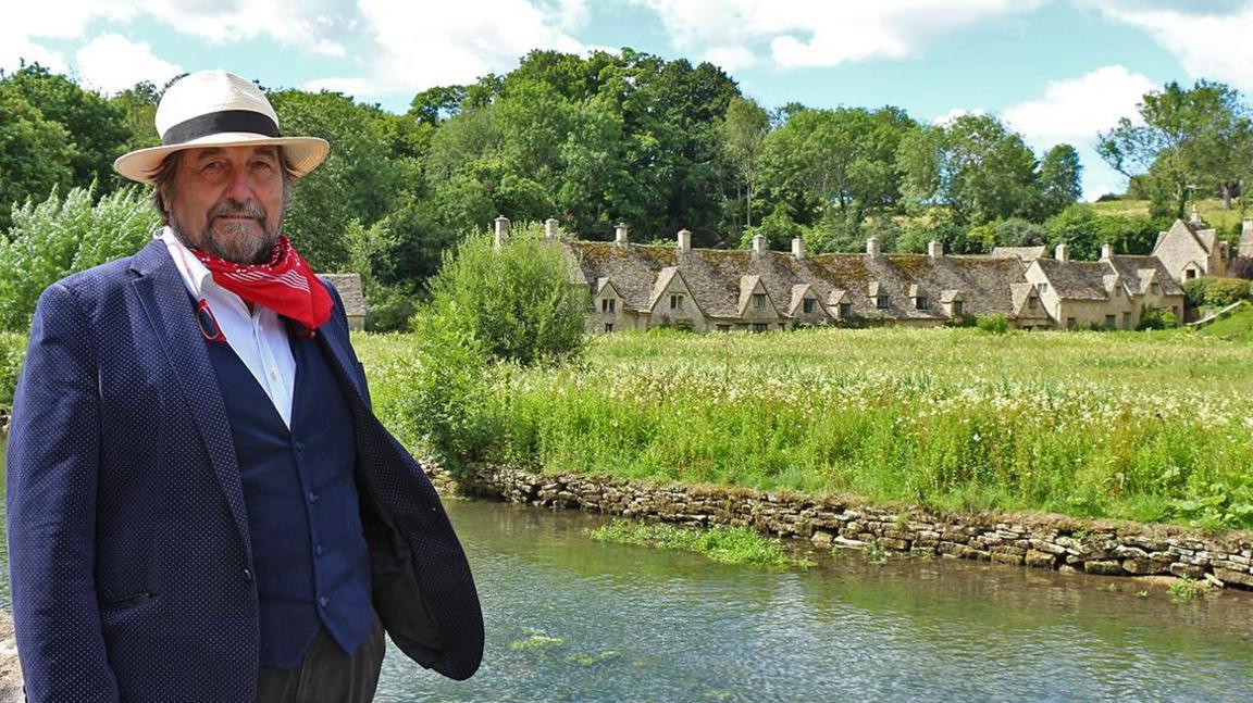 Mr Shuckburgh by a river on a sunny day. He is looking at the camera. There are old cottages on the other side of the bank, on a green meadow.