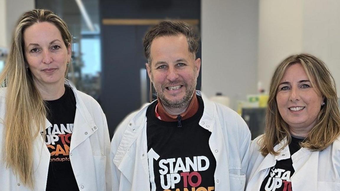 Left to right: Prof Petra Hamerlik, Alex and Emma Davies in a lab in the Manchester Cancer Research Centre. They are all wearing white coats and smiling. 