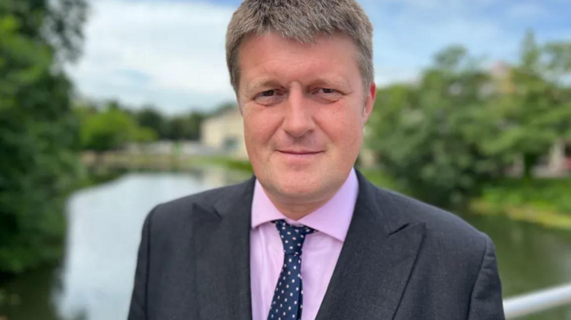 Councillor Richard Clewer standing in front of a body of water wearing a suit