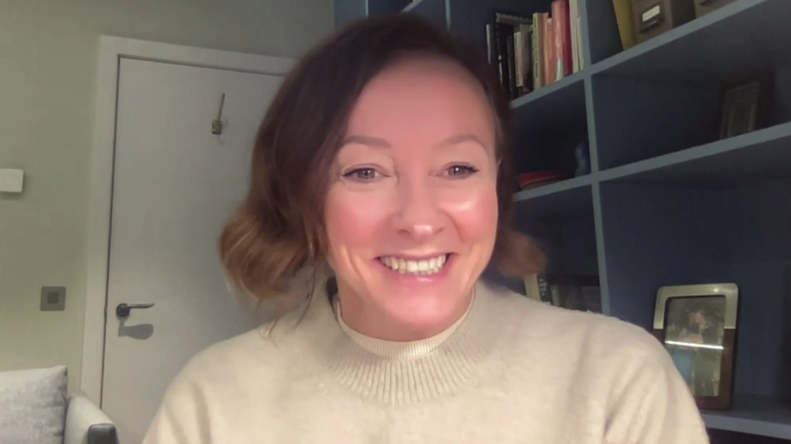 A woman, Mimi Turtle, wearing a cream coloured jumper. She has brown shoulder length hair, and behind her are blue coloured bookshelves.