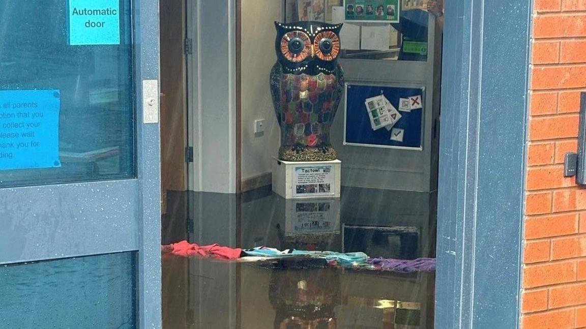 Water on the floor of a school room with multi-coloured cloth on the floor and a statue of an owl standing in a pool