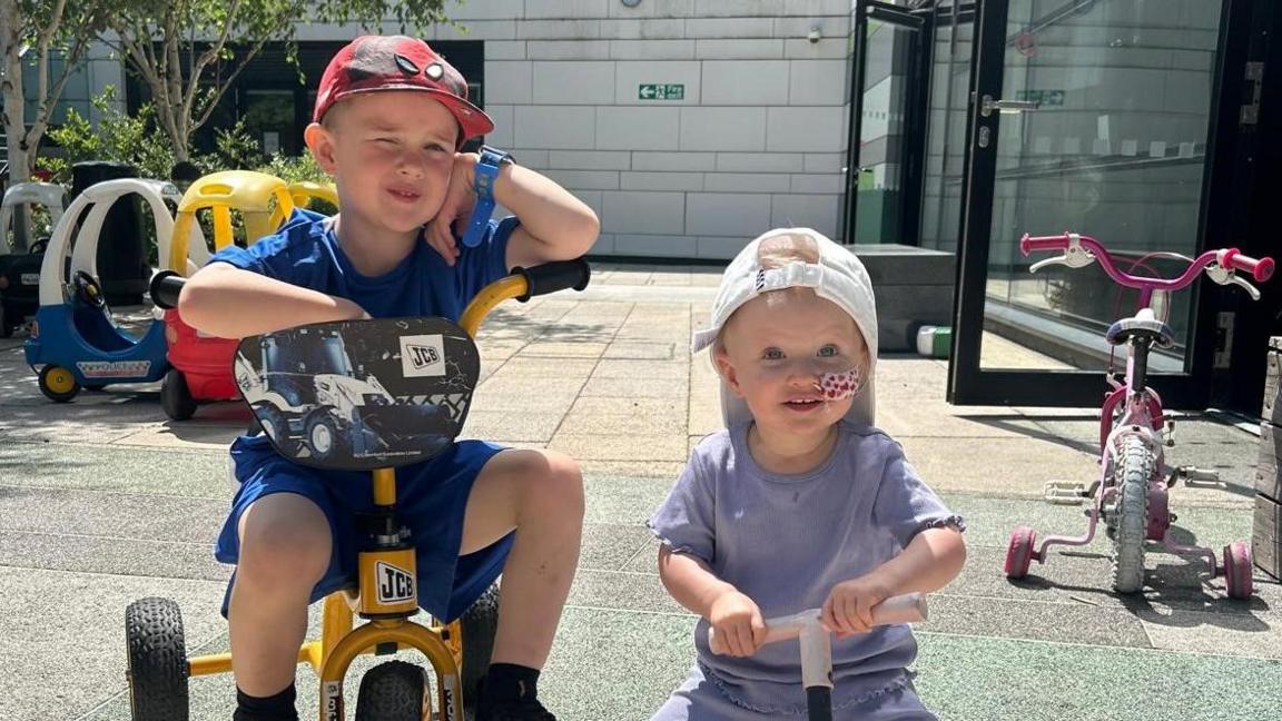 A four-year-old boy wearing a Spiderman cap sits on a trike with his face leaning against his hand. A little girl is on a scooter next to him. She has a feeding tube in her nose and is smiling.