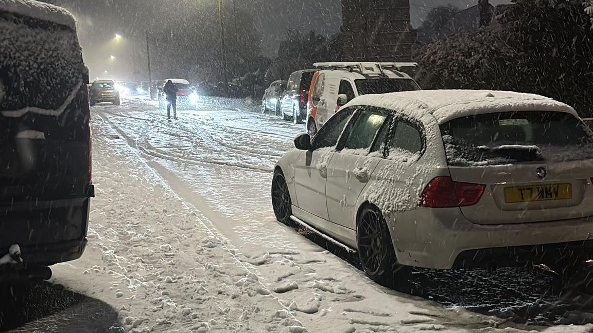 A white car on a road covered in snow. There are other cars on either side of the road. 