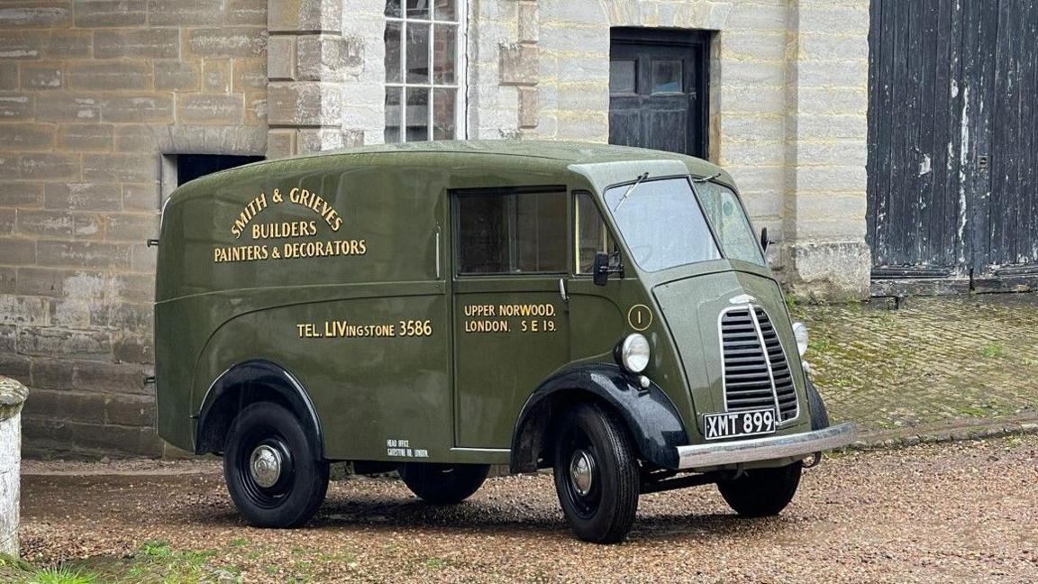 An olive green-coloured original Morris JE van is parked up on gravel in front of a stone building. It has 'Smith & Grieves Builders Painters & Decorators' painted on the side, along with 'Tel: Livingstone 3586' and 'Upper Norwood. London. S E 19'.  