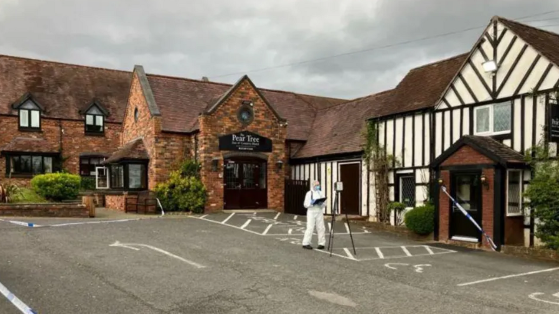 The Pear Tree Inn. A red brick building with an officer is forensic PPE and police tape sealing off a black door into the building.