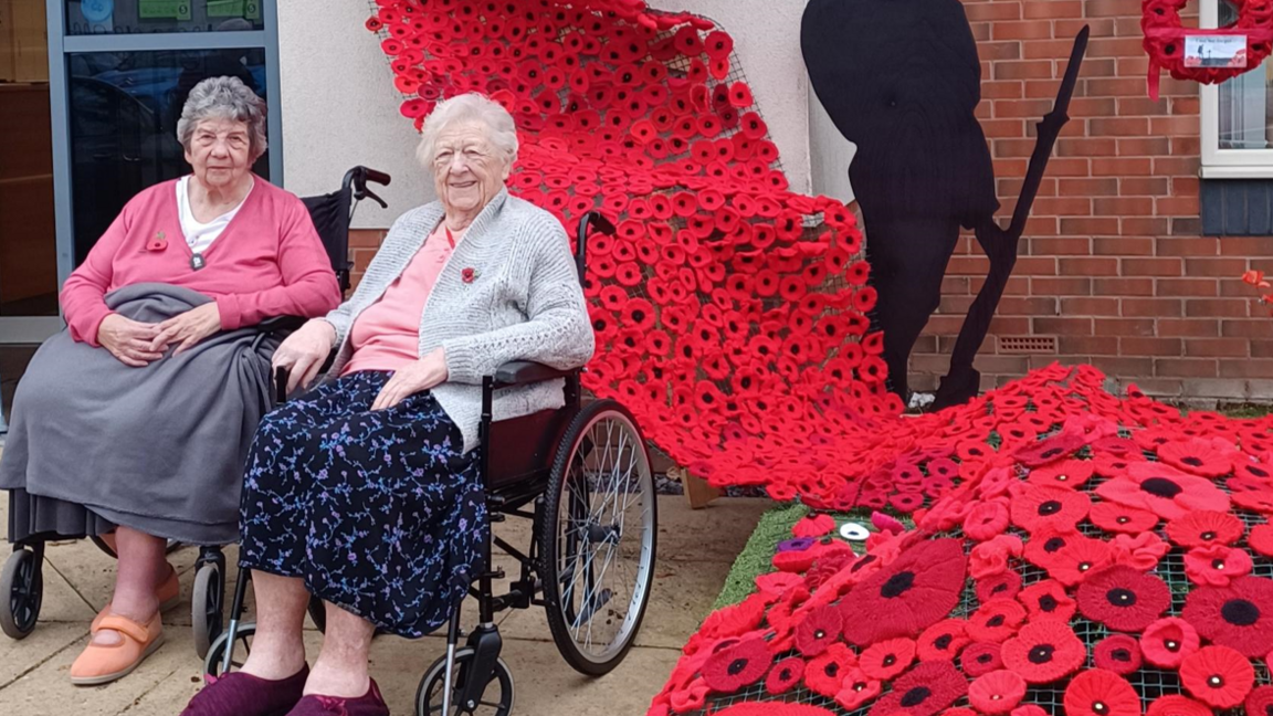 A stream of poppies tied to the side of a building's entrance with two ladies in wheelchairs sat to the left hand side