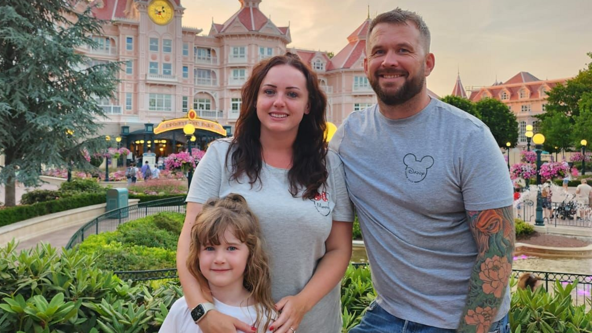 Angharad Dennis and husband Joe are wearing grey t-shirts with the Mickey Mouse logo. They stand in a park at Disneyland smiling with their daughter Cari.