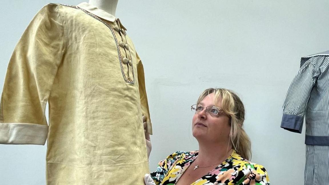 A woman wearing glasses and a floral dress examines a gold-coloured shirt