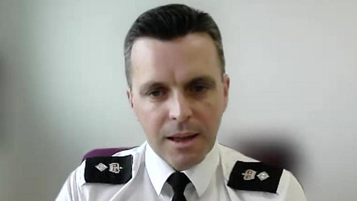 Acting Ch Supt Antony Hart of Devon and Cornwall Police speaks while on a Zoom call. His background has been blurred. He has short black hair which is greying around the side. He has a white shirt with black epilates on and a black tie.