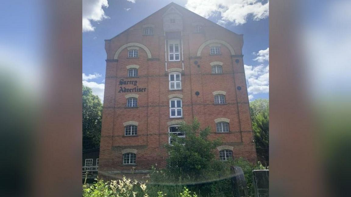 The outside of the former Surrey Advertiser building, which has the name of the paper on the front in black lettering. It is a six-storey mill building in red brick with windows all the way up and greenery growing around the ground floor.