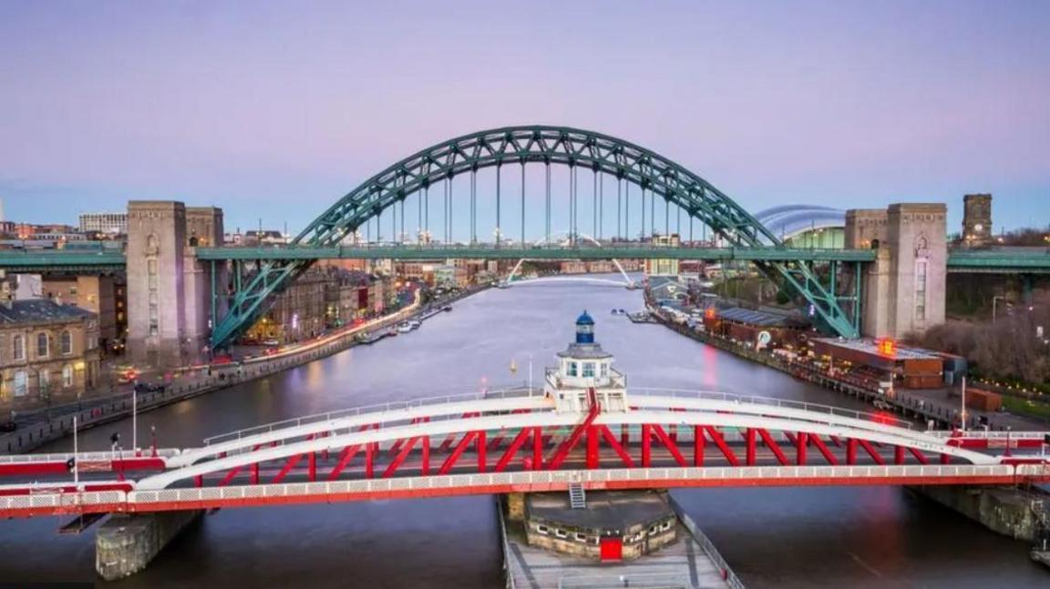 Aerial view of the Swing Bridge spanning the River Tyne - the structure's red and white metal girders rise to a low curve in the centre. Behind it, and down river can be seen the Tyne Bridge - a high metal arch with concrete pillars, and behind that the white curve of the Millennium footbridge.