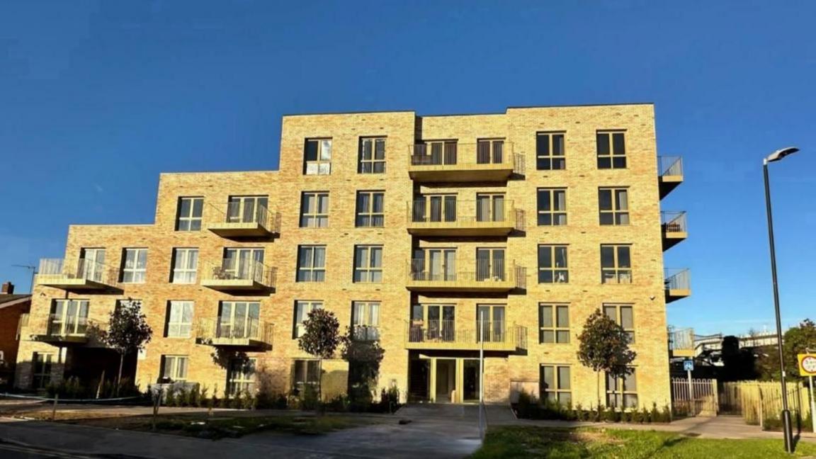 A modern yellow brick five-storey building with balconies to the front and sides and a concrete pathway leads to a main entrance at the front. Three small trees stand in front of the building