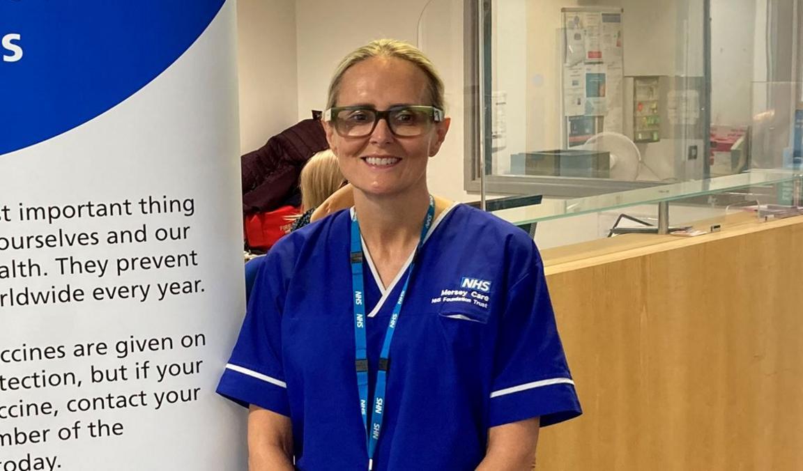 Julie Connell, who has tied-back blonde hair, wears her blue NHS Mersey Care NHS Foundation Trust uniform. She is wearing dark framed glasses and an NHS lanyard and is smiling for the camera.