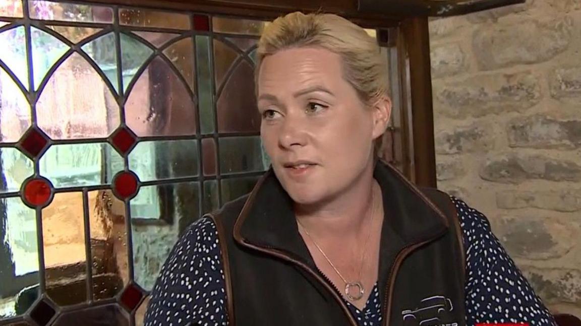 Woman with blonde hair wearing a speckled top and gilet sitting at a table in the pub with ornate window in the background