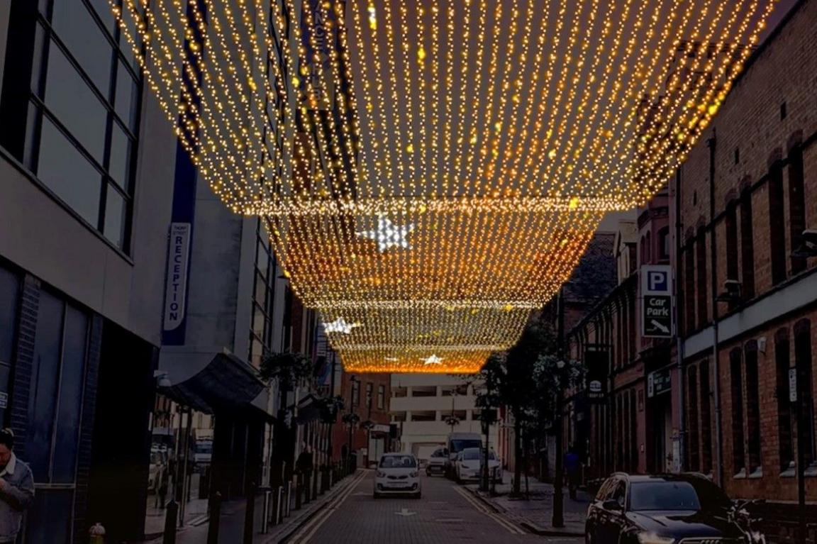 Many yellow lights hung above Thorp Street. Some cars are parked on the right hand side of the photo. There are buildings to the left and right of the road.