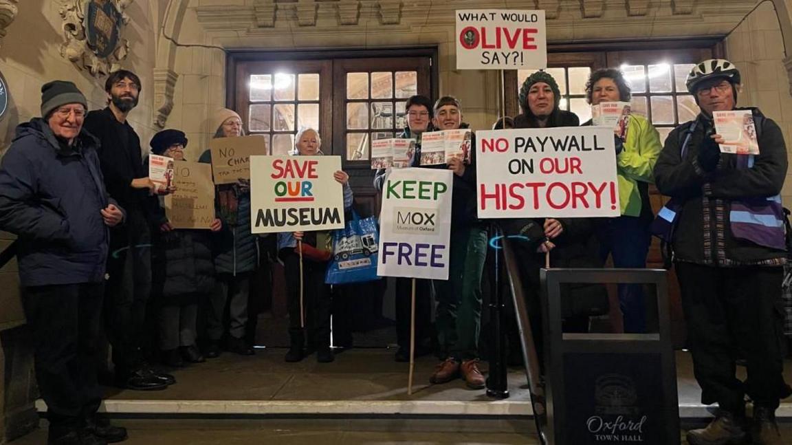 Ten protesters gather at the top of the entrance steps of the town hall. Placards include 'save our museum' and 'no paywall on our history'.