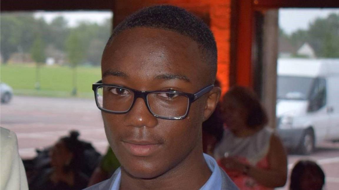 An image of Jamel Boyce, a teenage boy with very short dark hair and black-rimmed glasses, wearing a grey suit and blue shirt. Other people, a car park, a field and a white van can be seen in the background