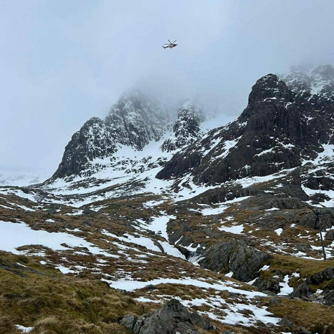 A coastguard helicopter flies over a mountain landscape covered in large patches of snow.