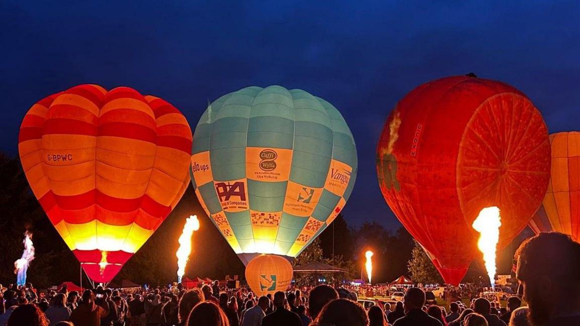 The image shows three entire balloons on the ground at night, with the backs of heads of people in the crowd visible at the bottom of the photo.