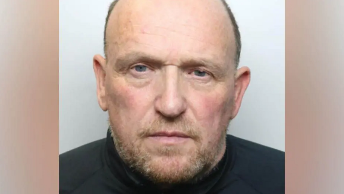 Police custody image of Nicholas Martin. He has receding short grey hair and brown and grey beard and is wearing a black top.