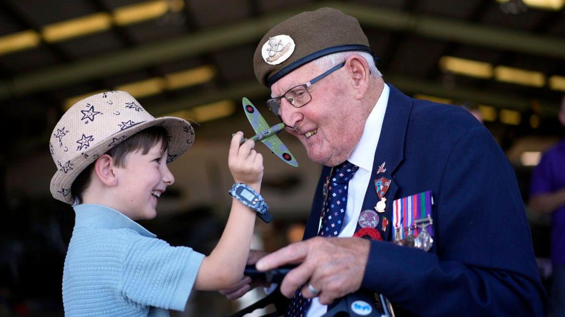 World War Two veteran Ivan Staines holds a small model Spitfire while standing beside five-year-old Alfie Rose in an aircraft hangar. Ivan Staines wears medals and a beret while Alfie Rose wears a hat and blue top.