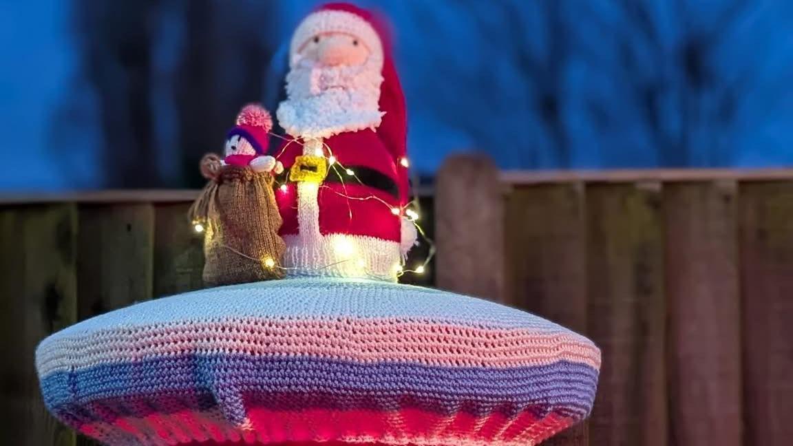 A purple and pink topper by the Red Lion in Wellington, Shropshire, is decorated with a Father Christmas holding a sack of presents with a toy soldier peeping out of the top. The display is decorated with white fairy lights.