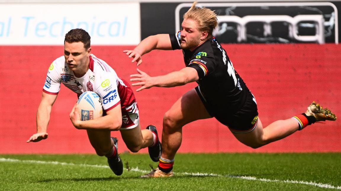 Wigan Warriors full-back Jai Field, in white and red, dives over the line to score a try as a Bradford player tries to tackle him