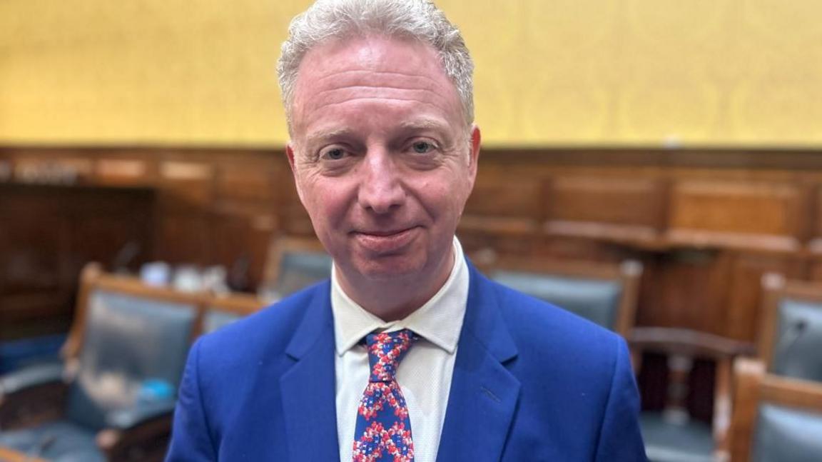 Alex Allinson standing in the Tynwald chamber looking straight ahead smiling. He is wearing a blue suit with a white tie and a blue, white and red tie.