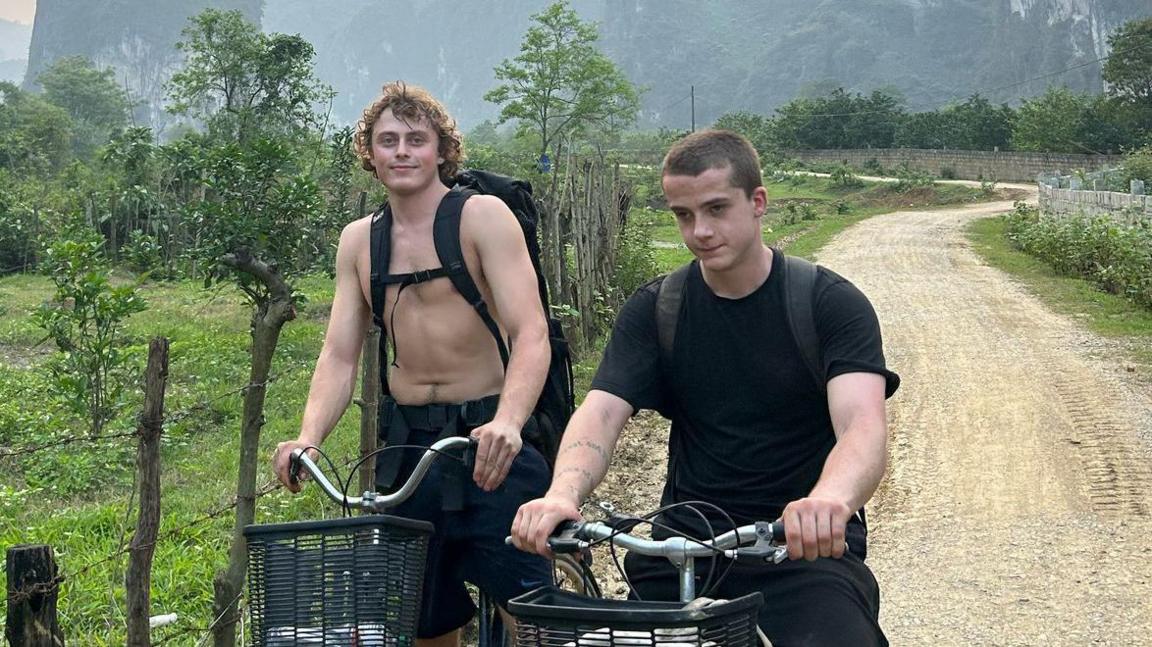 An image of Lux Losey-Sail (right), a young man with short shaved hair, wearing a black T-shirt and sitting on a bike next to his brother Max (left), who is also sitting on a bike, on a dirt track with plants and mountains behind them