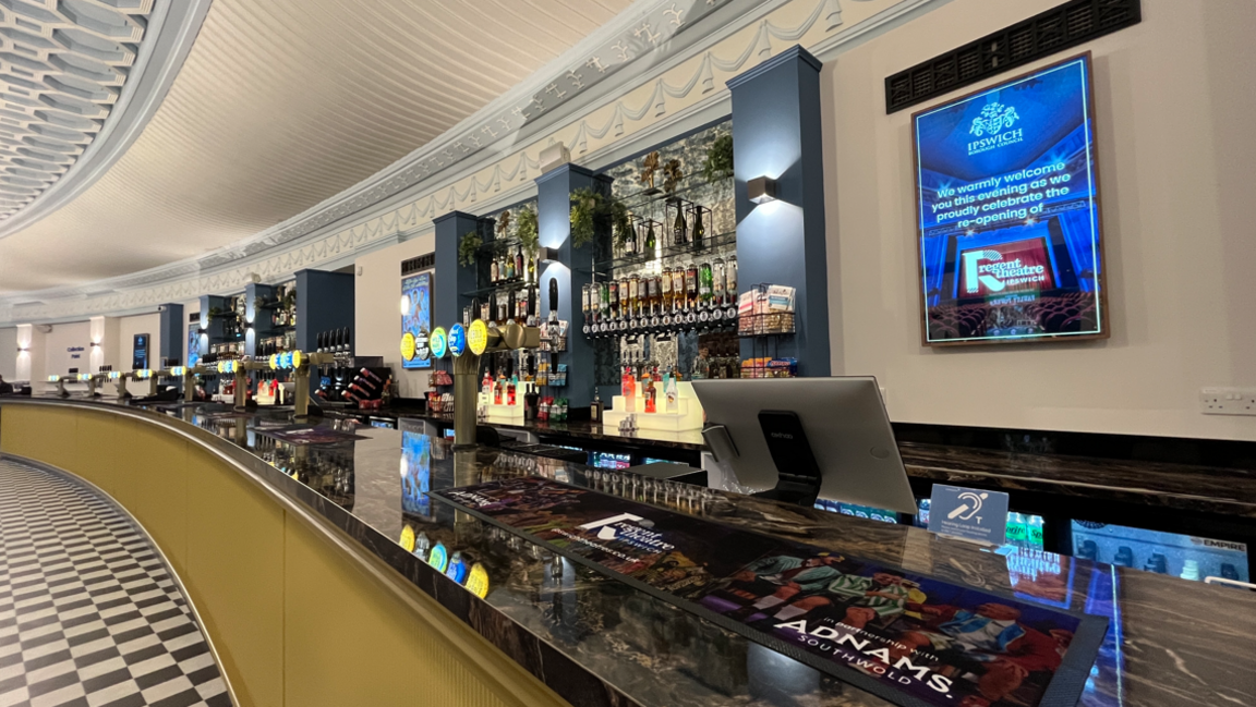 A view of a long bar, showing beer pumps, optics, and a beer mat in the foreground with Adnams written on it. The floor is a black and white check.