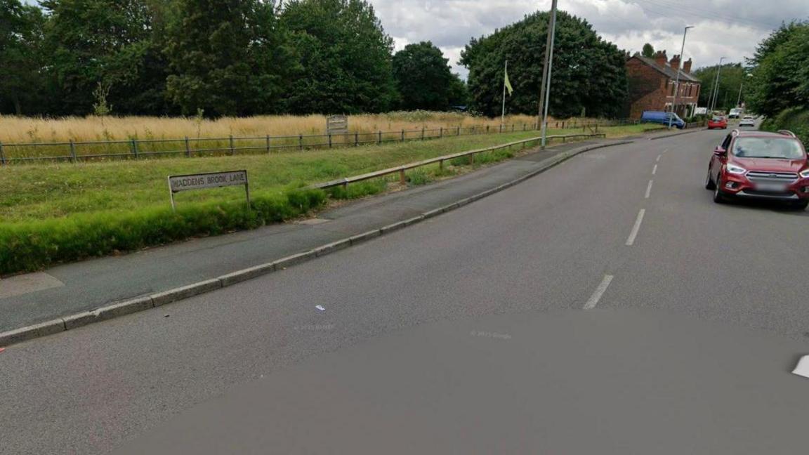 The start of a road with Waddens Brook Lane sign on the left hand side near a grass verge. On the right hand side is a red car driving out Waddens Brook Lane. House in the distance with a blue van parked outside.
