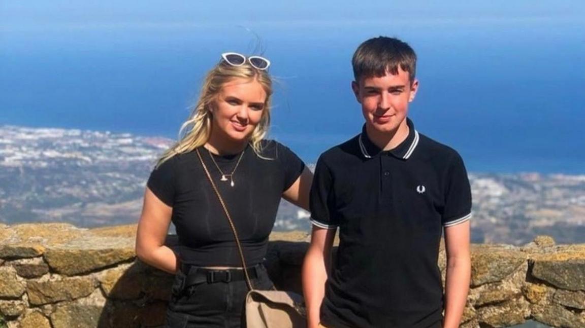 A young woman with long blonde hair dressed in black stood beside a young man with short brown hair wearing a black Fred Perry polo top. They are looking into the camera and smiling while stood with their backs facing what appears to be a viewing point above a coastline.