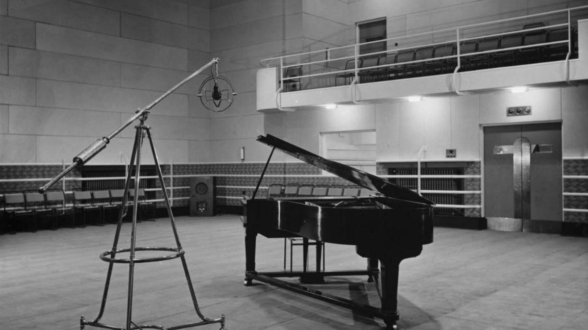 A black and white image of a recording studio. It is a fairly blank room with a grand piano sitting in the middle and a microphone on a stand poised above the piano. There is also an overhead seating area to the right.