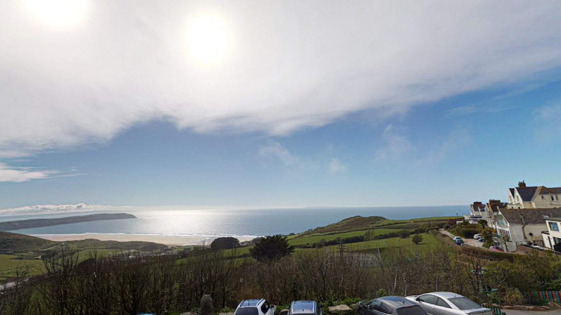 A scenic coastal photo looking out from a hotel balcony with the sea in the distance. Bright blue skies with the blue sea below. There is lots of green fields as well as some buildings to the right hand side of the photo. There are also a few cars parked and a sandy beach in the background.