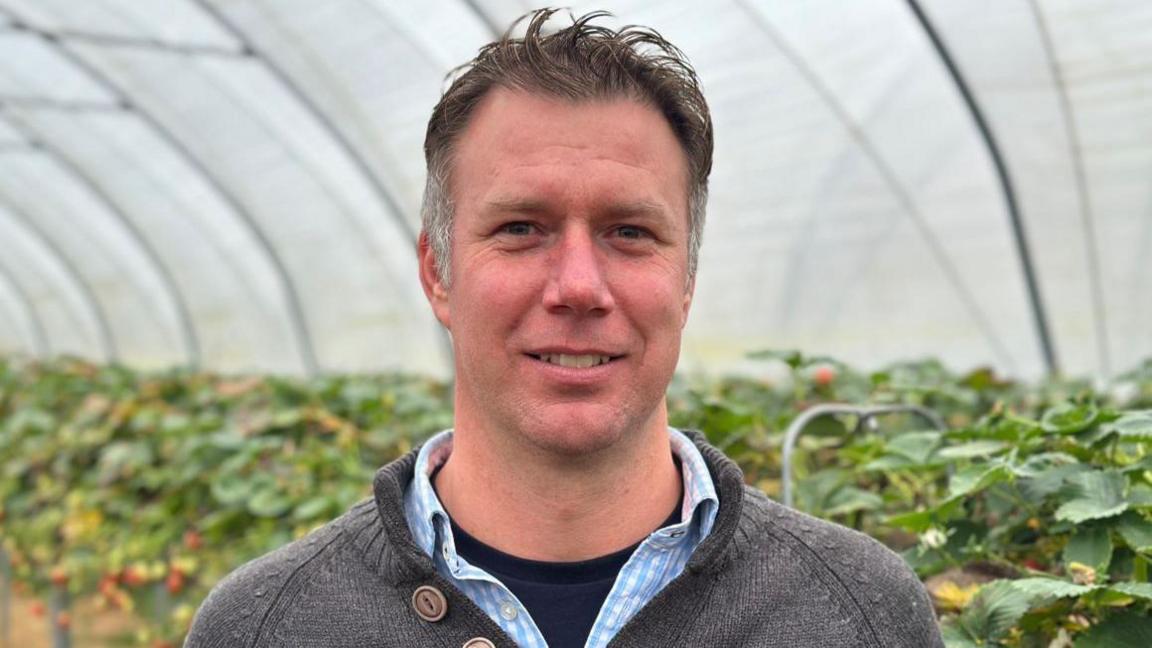 A man with dark hair and a grey top in front of a row of plants