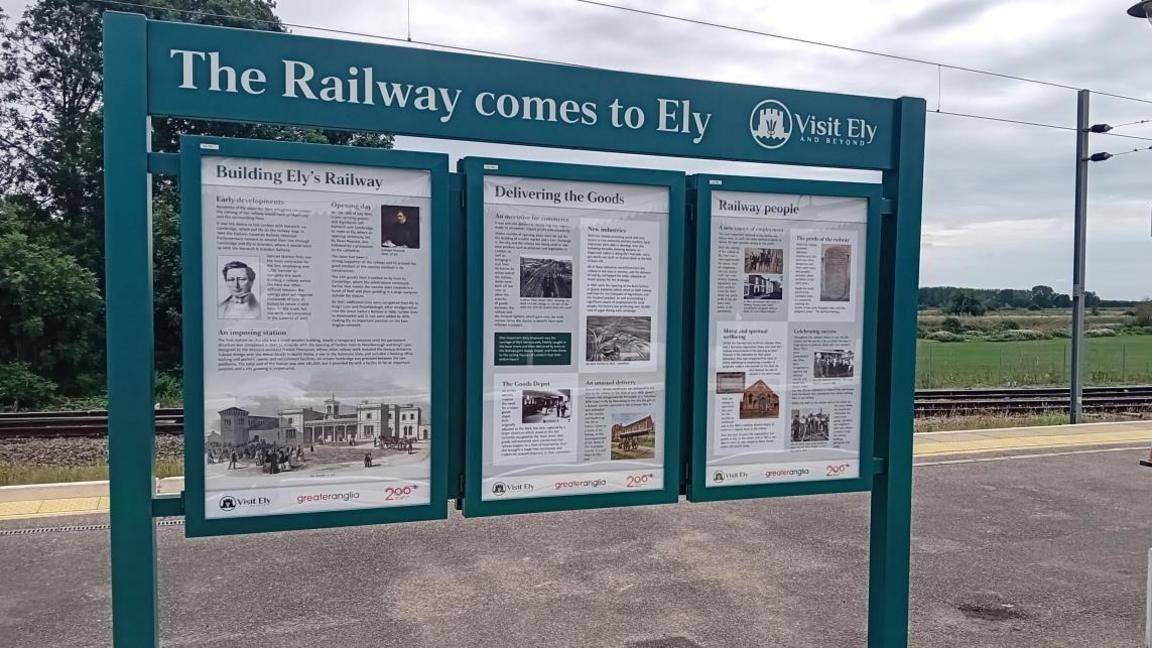 Three public notices which are history boards in dark grey set up on the platform of the railway station, they show various images and text.
