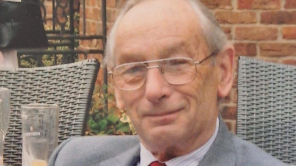 An elderly man with thinning grey hair and glasses smiles at the camera. He sits in front of a red brick wall with a glass visible to his right and the backs of rattan chairs.