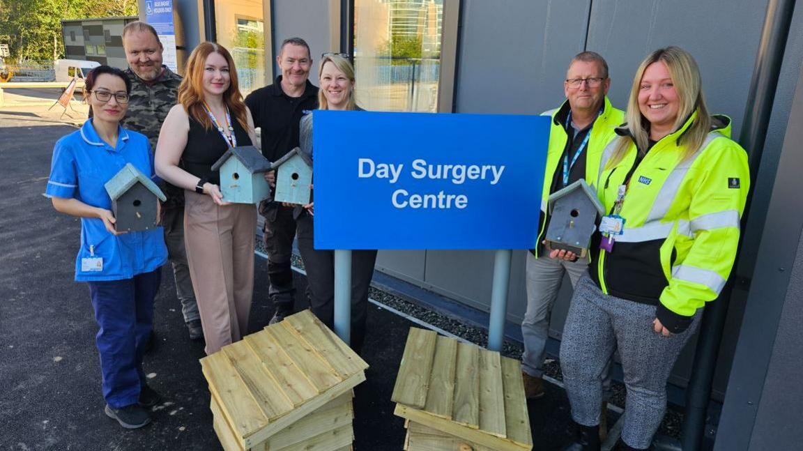 Tripti Thapa, day surgery staff nurse; Jim Hicks, Worklink service user; Chloe Michaels, day surgery team leader; Graham Willis, employment support trainer, Worklink; Abby Gibbon, senior employment officer, Worklink; Mike Todd, NHS project manager; Amy Lockyer, NHS commissioning and support services manager. Holding blue bird boxes in front of the day surgery centre with two larger wooden boxes on the ground