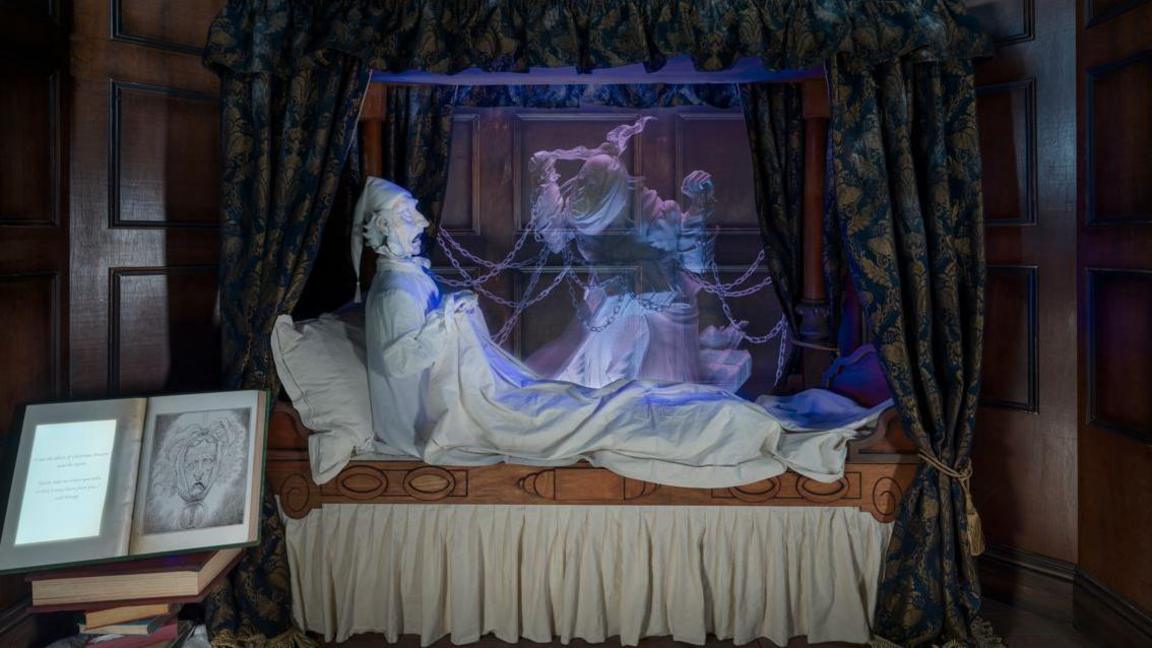 A tableau depicting a scene from A Christmas Carol. The figure of Scrooge is sitting in a four poster bed and it confronted by the ghost of his business partner - a semi transparent figure draped in chains. An opened book with an illustration can be seen to the left.
