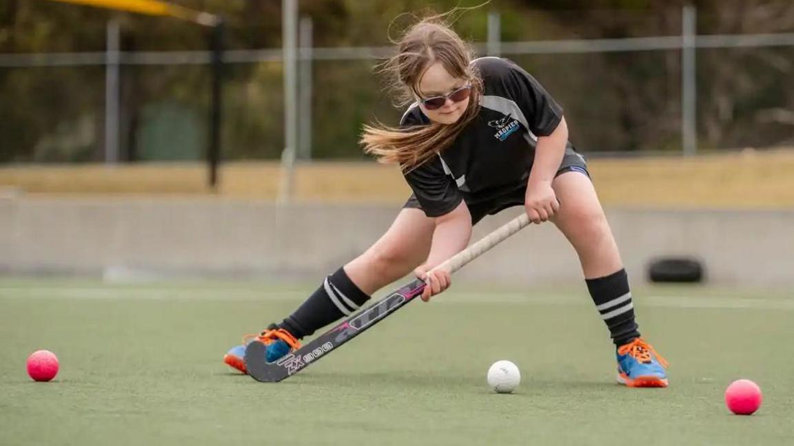 A girl wearing black sports shorts and t-shirt leans forward to hit a white ball with a hockey stick