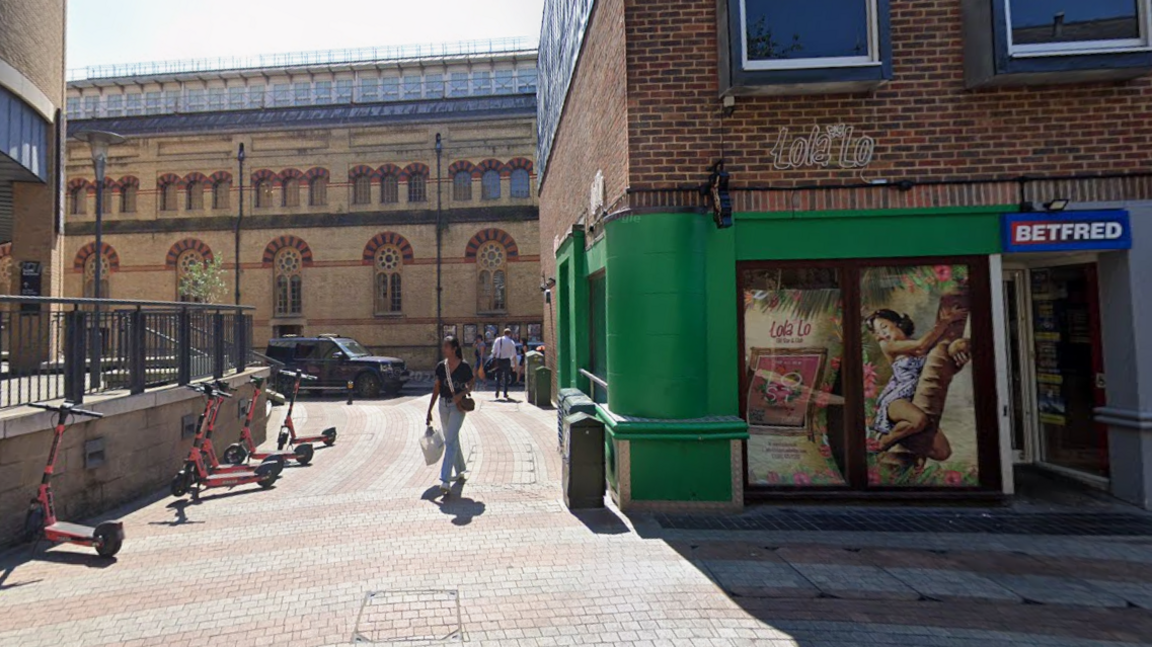 An external image of Lola Lo nightclub in Cambridge. It is on the corner of a brick-built building with a green door and window frames. Above it says Lola Lo. The entrance to the neighbouring Bedfred bookmakers unit is visible.