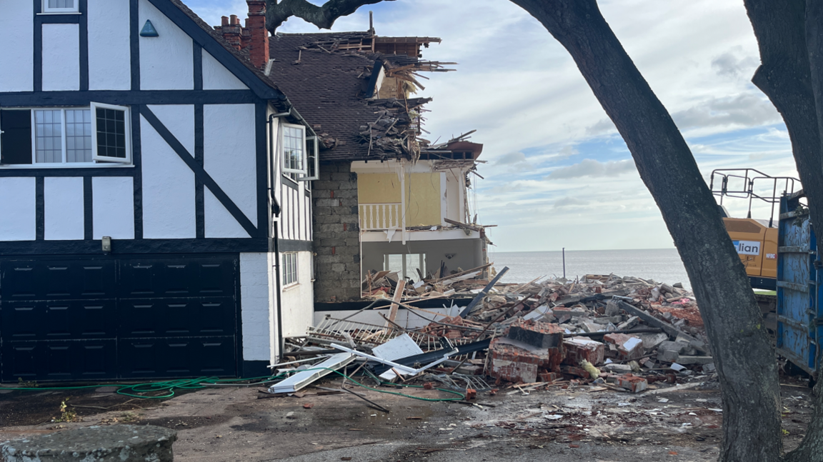 A partially demolished home on the coastline next to a pile of debris. In the background is the sea, blue sky and white clouds.